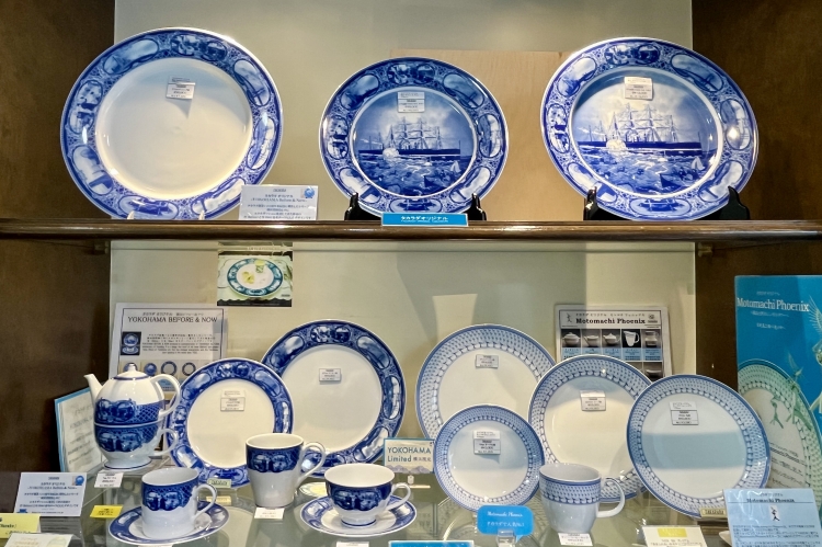 Una serie original de platos decorativos, tazas y otros artículos que presentan paisajes de Yokohama representados en un vibrante color azul.